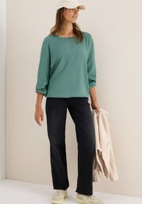 Vrouw in een groene blouse met lange mouwen, zwarte broek, witte sneakers en een witte pet, die een beige jas in haar rechterhand houdt.