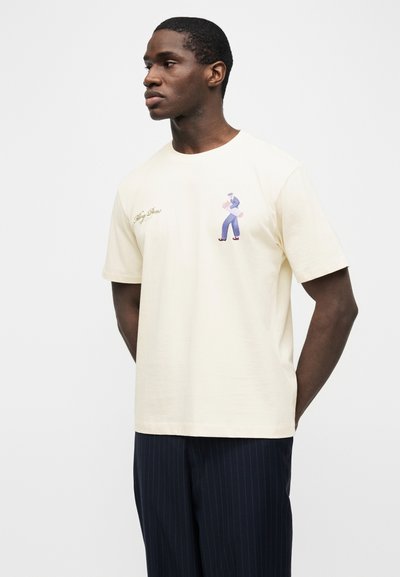 Άνδρας που φορά κρεμ κοντομάνικη μπλούζα με μικρό σχέδιο ενός πεζοπόρου και γραφικό κείμενο σε καλλιγραφία στο αριστερό στήθος, συνδυασμένη με σκούρο παντελόνι με λεπτές ρίγες.