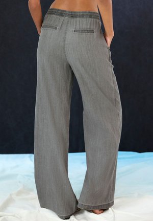Person wearing loose gray pants with elastic waistband, hands in side pockets, standing barefoot on a white fabric surface.