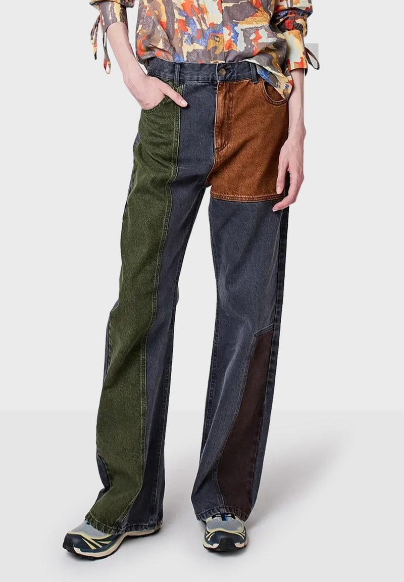 Person som bär vida patchworkjeans med gröna, mörkgrå, bruna och svarta denimsektioner, tillsammans med sneakers och en färgglad skjorta.