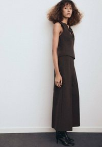Haut sans manches marron associé à un pantalon large, avec un détail de nœud au niveau du col, fabriqué en tissu lisse. Porté avec des bottines noires à la cheville.