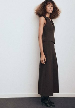 Haut sans manches marron associé à un pantalon large, avec un détail de nœud au niveau du col, fabriqué en tissu lisse. Porté avec des bottines noires à la cheville.