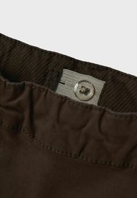 Brunt stoff med et teksturert midjebånd, med en lysegrå stoffetikett og en gjennomsiktig knappelukking. Synlige sømdetaljer.