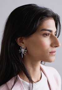 Orecchini in argento a forma di ragno stilizzato, con dettagli intricati e una superficie lucida e strutturata, indossati sull'orecchio di un modello.