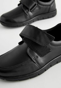 Chaussures en cuir noir avec une bande Velcro, bout rond, dessus texturé et semelle en caoutchouc avec des détails striés pour une meilleure adhérence.