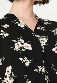 Primo piano di un indumento nero a V con motivi floreali bianchi e rosa indossato da una persona con i capelli corti castani.