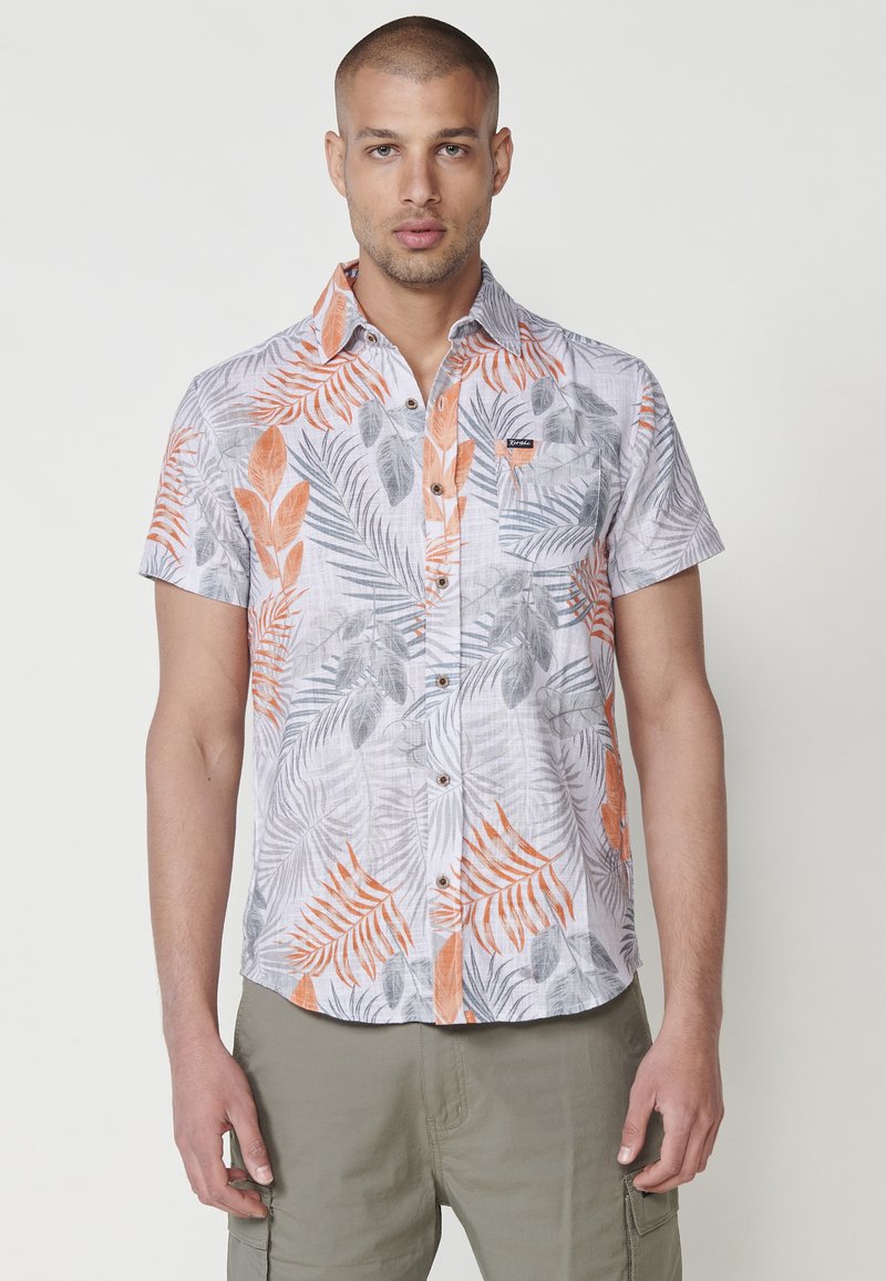 Cameriera a maniche corte in tessuto leggero, con una stampa di foglie tropicali arancioni e grigie, dotata di tasca e branding discreto.
