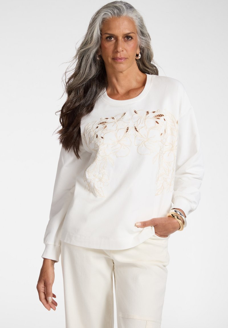 Donna matura con lunghi capelli grigi che indossa una felpa bianca ricamata con fiori, pantaloni bianchi, orecchini a cerchio e numerosi braccialetti, posando con sicurezza.