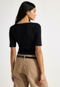 Musta ribbineulottu lyhythihainen toppi yhdistettynä korkeavyötäröisiin beigen värisiin housuihin. Asussa on musta vyö ja housujen takana taskut.