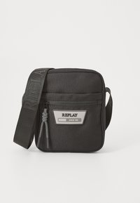 Black fabric crossbody bag with adjustable strap, zip closure, front logo patch, and woven accent details. Compact rectangular shape.