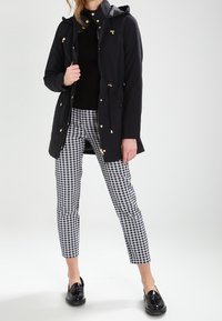 Svart huva jacka med guldfärgade detaljer, bärs över en åtsittande svart topp och svart-vitt rutig byxor. Svarta skor kompletterar outfiten.