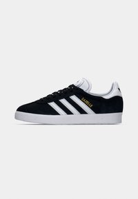 Svart Adidas Gazelle-sneakers med hvite striper, hvit såle og gullfarget "GAZELLE"-tekst på siden, vist i profil mot en enkel bakgrunn.