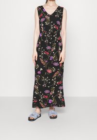 Svart blommig maxiklänning med V-ringning, med livfulla mönster av lila och röda blommor. Bärs med ljusblå sandaler.