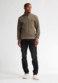 Jasnobrązowa kurtka zapinana na zamek z teksturowanej tkaniny, z prążkowanymi mankietami i dołem. Ubrana na ciemne dżinsy i jasne trampki, stoi na jednolitym tle.