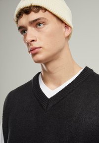 Black knitted sweater with a v-neck design layered over a white undershirt, paired with a cream-colored knit beanie. Smooth texture.