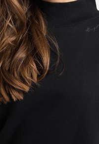 Musta poolopaita pehmeästä kankaasta, jossa on resorikaulus. Yksityiskohtina hienovarainen kirjailu kaula-aukossa ja löysät, aaltoilevat ruskeat hiukset.