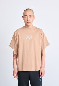 Bež veliki T-shirt s okruglim izrezom i kratkim rukavima. Ima bijeli Nike logo otisnut na prsima. Meka tkanina.