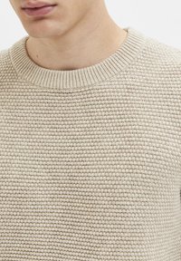 Beiger Strickpullover mit rundem, geripptem Ausschnitt. Verfügt über ein strukturiertes Muster und einen weichen, leicht elastischen Stoff.