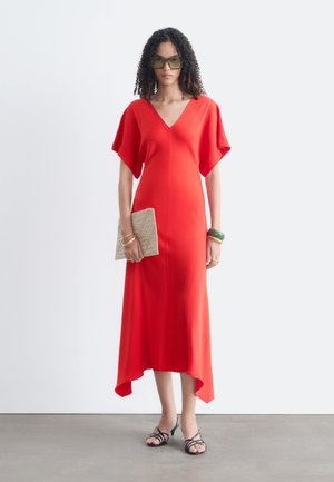 Femme aux cheveux bouclés portant des lunettes de soleil vertes et une robe midi rouge, tenant une pochette beige, portant des bracelets et des sandales noires.