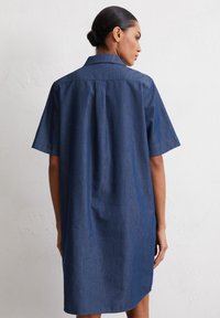 Denim jurk in donkerblauw met korte mouwen, een puntige kraag en een rugplooien voor extra bewegingsvrijheid. Rechte snit tot op de dij.