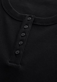 Top in tessuto nero a coste con scollo rotondo, chiusura frontale con cinque bottoni neri e superficie strutturata.