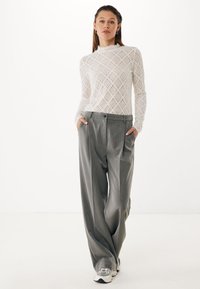 Top bianco in pizzo a maniche lunghe con motivo a rombi, abbinato a pantaloni grigi a gamba larga, con pieghe frontali e chiusura con bottone, indossato con sneakers.