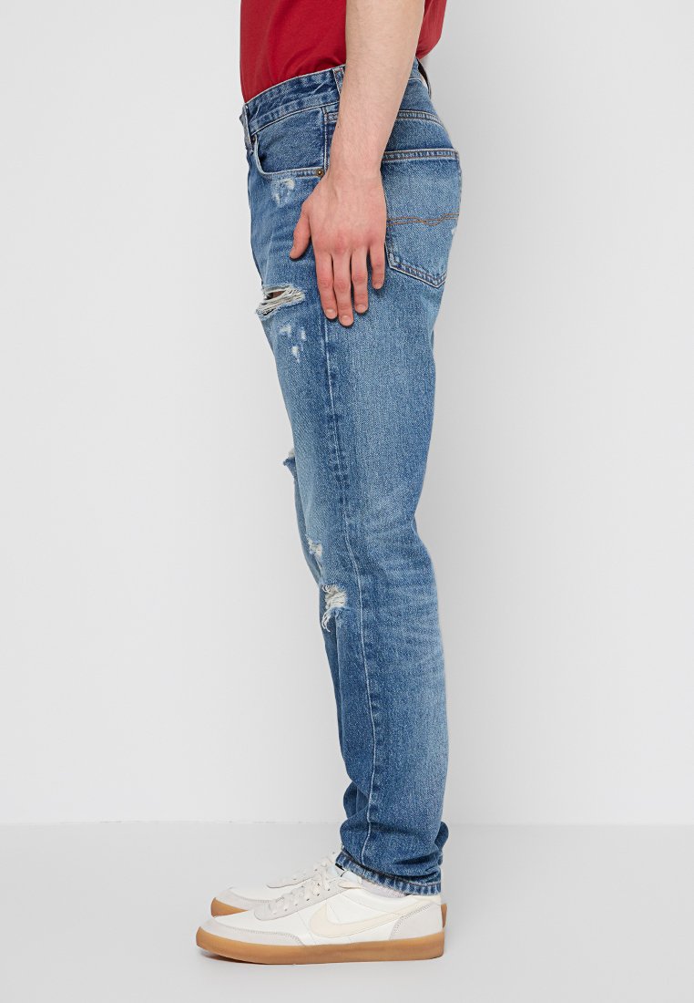 Schlanke, blaue Denim-Jeans mit verwaschenen Details. Verfügt über fünf Taschen und helle Nähte. Getragen mit weißen Sneakers.