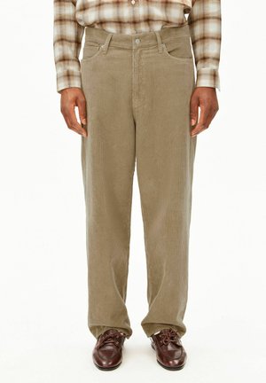 Ein Mann trägt beige Cordhosen, braune Leder-Schnürschuhe und ein langärmeliges kariertes Hemd und steht vor einem weißen Hintergrund.