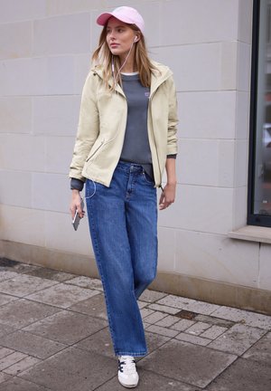 Junge Frau, die im Freien spazieren geht, trägt eine beige Jacke, blaue Jeans, eine rosa Kappe, weiße Turnschuhe und hört Musik mit Ohrhörern, die an ihr Telefon angeschlossen sind.