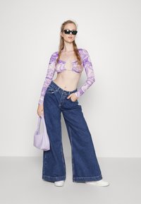 Camisola roxa de mangas longas com um padrão marmoreado, combinada com jeans de corte solto e ténis brancos. Uma mala de ombro lilás ilumina o visual.