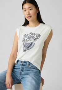 Biała koszulka bez rękawów z graficznym nadrukiem "Levi Strauss & Co." w kolorze niebieskim, w zestawie z jasnoniebieskimi dżinsami.