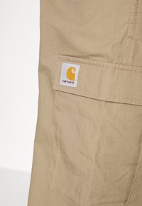 Beigenvärinen kangasrunko, jossa valkoinen ja keltainen Carhartt-logoplässi. Materiaalissa on hienovarainen ruutukuvioinen tekstuuri.