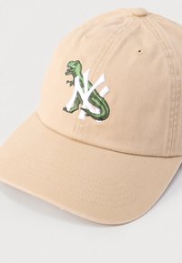 Μπεζ καπέλο μπέιζμπολ με κυρτό γείσο, που απεικονίζει ένα πράσινο δεινόσαυρο και λευκό κεντητό λογότυπο "NY"  στο μπροστινό μέρος. Υφασμάτινη υφή.