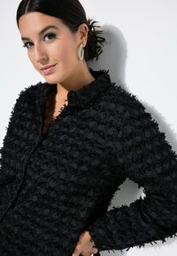 Maglietta nera in tessuto testurizzato con intricate fantasie in rilievo. Design con bottoni e colletto, con dettagli frangiati.