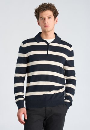 Junger Mann mit lockigem Haar, der ein dunkelblau-weiß horizontal gestreiftes Poloshirt mit langen Ärmeln und schwarze Hose trägt, steht vor einem einfarbigen Hintergrund.