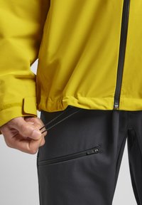 Giacca impermeabile gialla con chiusura a zip, orlo regolabile e pantaloni neri abbinati con tasche a zip e una texture liscia.