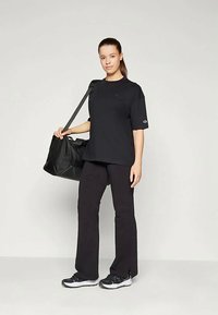 Mladá žena stojí, oblečená v čiernom oversized tričku, čiernych nohaviciach so bočnými rozparokmi, čiernych teniskách a nesie čiernu športovú tašku na pleci.