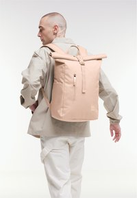 Rolgedeikte rugzak van zacht roze stof. Voorzien van gevoerde schouderbanden, een voorvak met ritssluiting en logo-details. Eenvoudig geometrisch ontwerp.