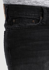 Primer plano de un bolsillo de jeans de mezclilla negra con costuras visibles, remaches y una camisa de patrón gris metida por dentro.