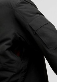 Zwarte buitenjack met een soepele textuur, schuine schoudernaden en een klepzak op de mouw. Binnenvoering in een contrasterende kleur.