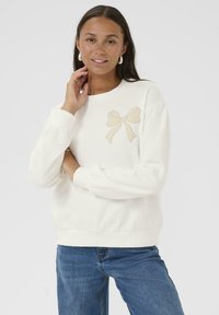 Sweatshirt blanc en matériau doux, doté d'un nœud beige clair brodé sur la poitrine gauche. Le design comprend des poignets et un ourlet côtelés.