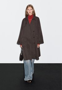 Długi ciemny płaszcz o strukturalnym kształcie, z dużymi guzikami i bocznymi kieszeniami, nałożony na czerwony golf. Noszony z jasnoniebieskimi dżinsami.