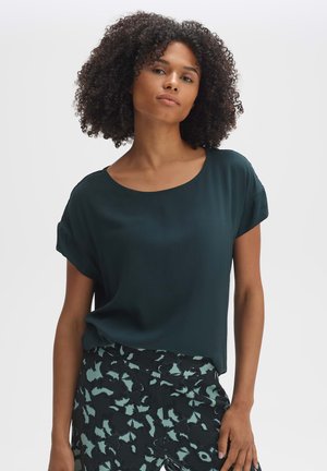 Blusa verde scuro a maniche corte con scollatura ampia, realizzata in tessuto morbido. Abbinata a pantaloni a fantasia neri e teal.