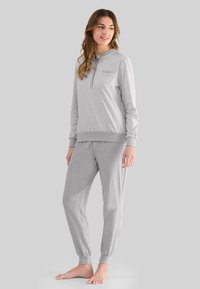 Mujer de pie descalza, vestida con una camiseta de manga larga rayada gris claro y pantalones con puños en los tobillos a juego, frente a un fondo gris liso.