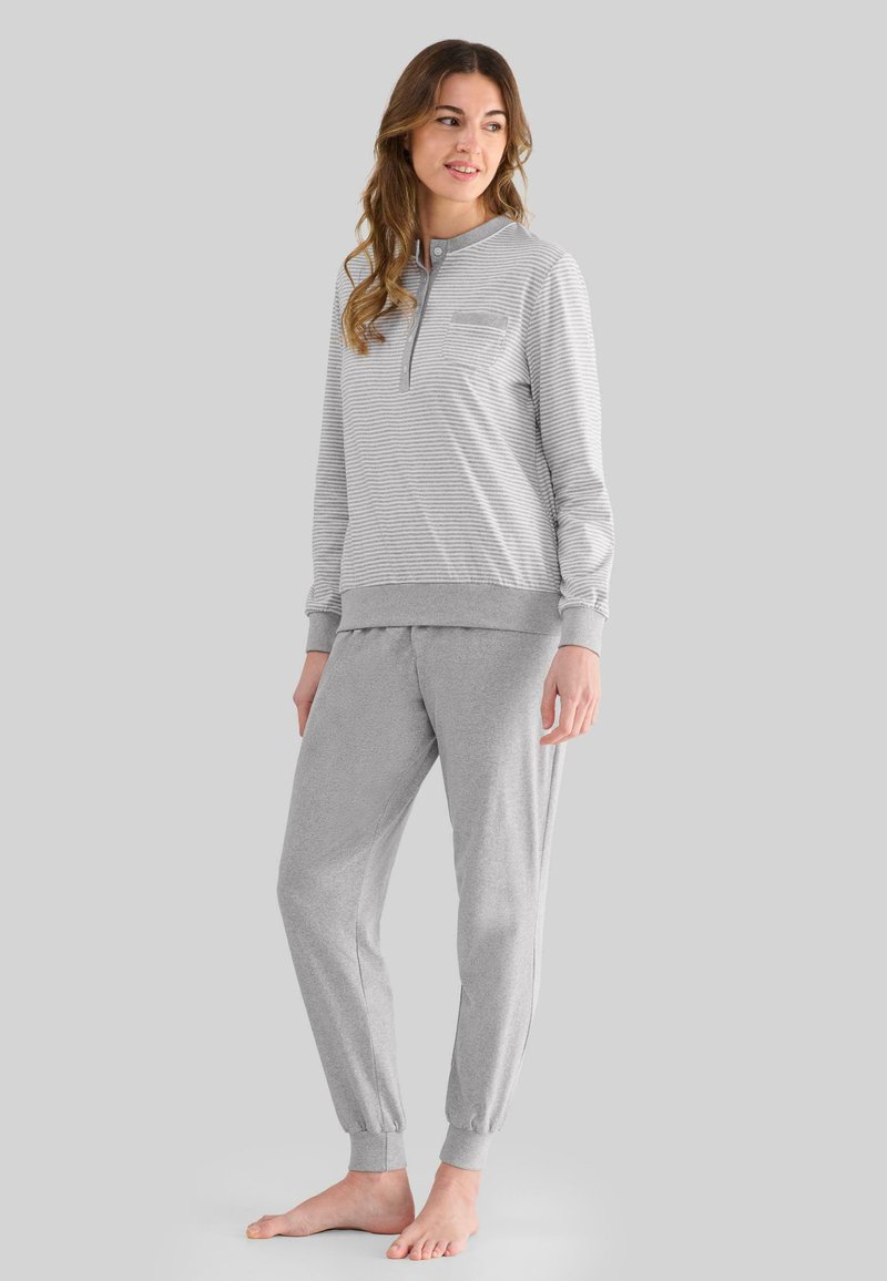 Mujer de pie descalza, vestida con una camiseta de manga larga rayada gris claro y pantalones con puños en los tobillos a juego, frente a un fondo gris liso.