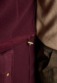 Ein burgunderfarbenes Fleece-Oberteil aus strukturiertem Stoff, ausgestattet mit einer goldenen Akzentnadel und einer Seitentasche, kombiniert mit einem beigen Pullover und gemusterten Hosen.