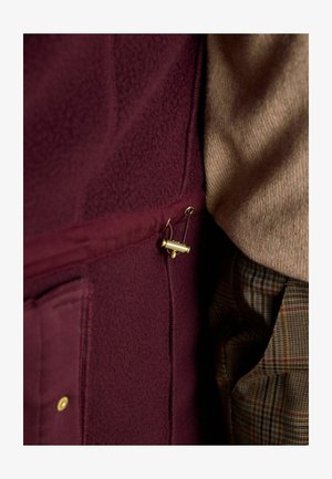 Ein burgunderfarbenes Fleece-Oberteil aus strukturiertem Stoff, ausgestattet mit einer goldenen Akzentnadel und einer Seitentasche, kombiniert mit einem beigen Pullover und gemusterten Hosen.