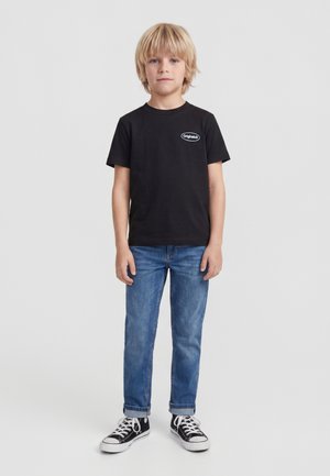 Blond jongetje staat, draagt een zwart t-shirt met het "Originals" logo, blauwe spijkerbroek en zwarte hoge sneakers met witte veters.