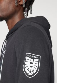 Schwarze Kapuzenjacke mit einer strukturierten Oberfläche, die ein auffälliges weißes Logo-Patch auf dem linken Ärmel und Zugbänder für die Kapuze aufweist.