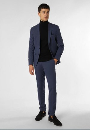 Männer-Anzug in schlanker Passform, blau mit Reverskragen, kombiniert mit einem schwarzen Rollkragenpullover und schwarzen Schuhen. Glattes Material, schmal zulaufende Hosen und umgeschlagene Säume.